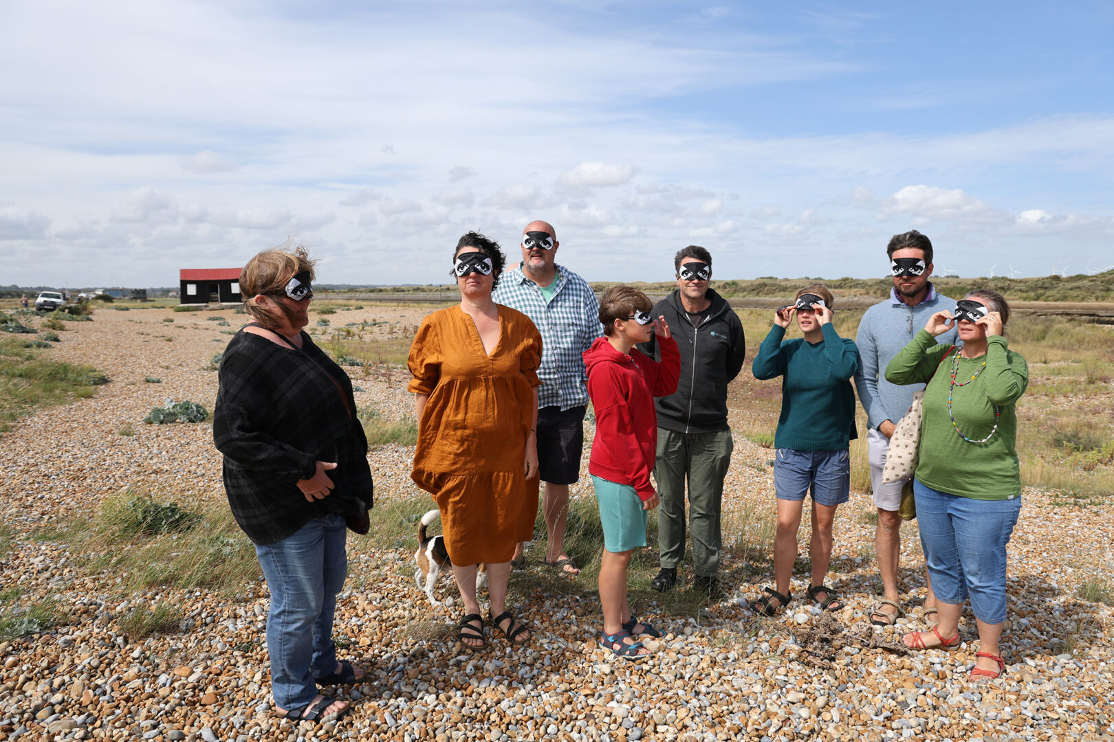 A group of people stand on a shingle beach wearing eye masks